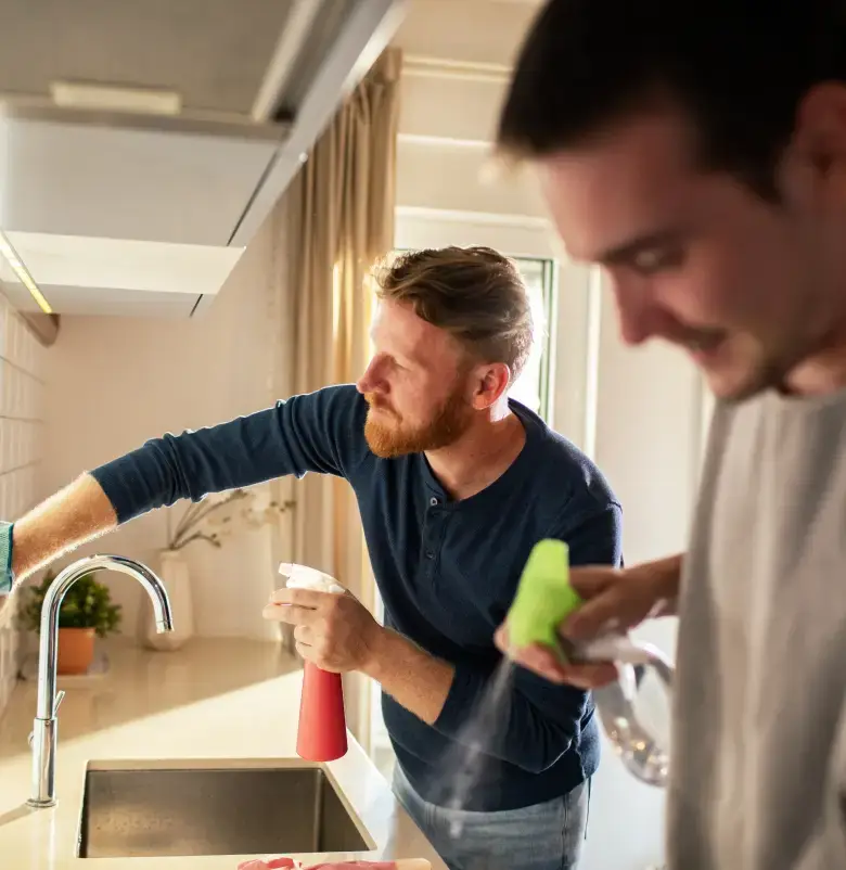 Two men cleaning a kitchen counter with spray bottles near a sink.